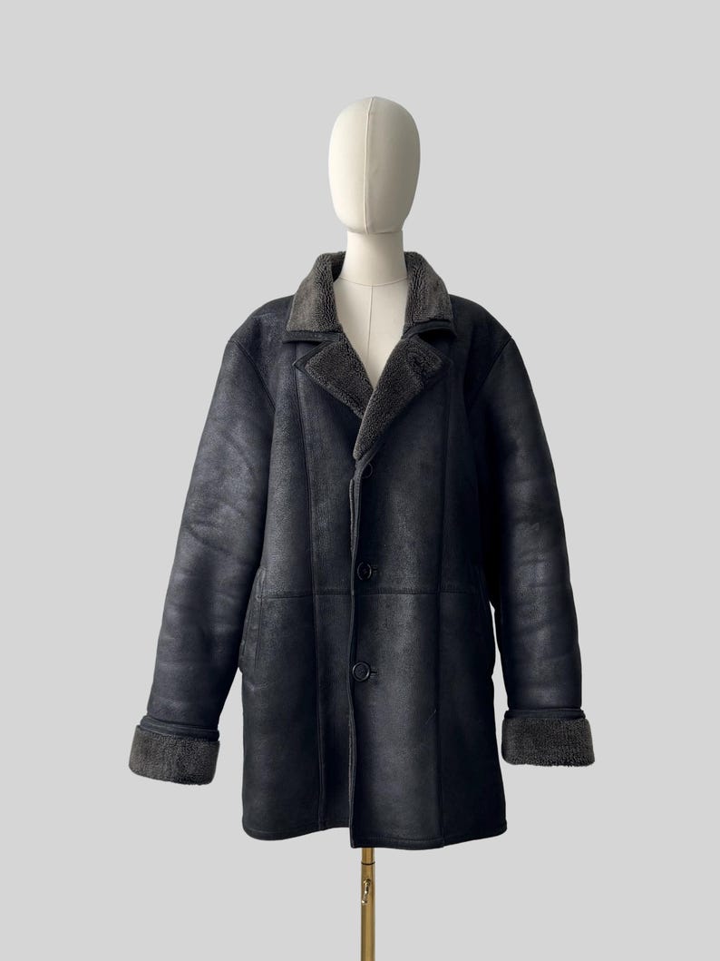 May include: A black shearling coat with a gray shearling collar and cuffs. The coat has a button-up front and is displayed on a mannequin. The coat is a classic style, suitable for cold weather.