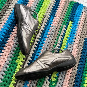 May include: A pair of silver leather slip-on shoes with black soles. The shoes are on a colorful crocheted rug.