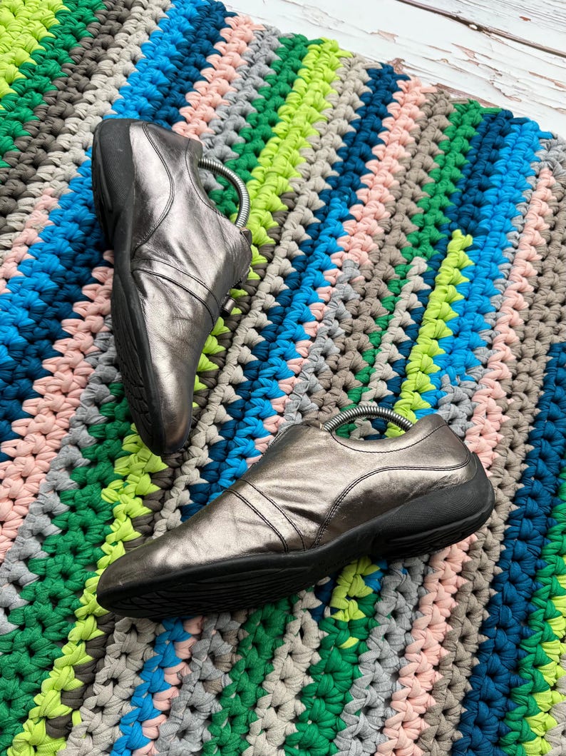 May include: A pair of silver leather slip-on shoes with black soles. The shoes are on a colorful crocheted rug.