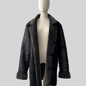 May include: A black shearling coat displayed on a mannequin. The coat features a dark gray, plush interior lining and collar, with matching cuffs. The exterior is a smooth, matte black. The coat has three dark buttons down the front.