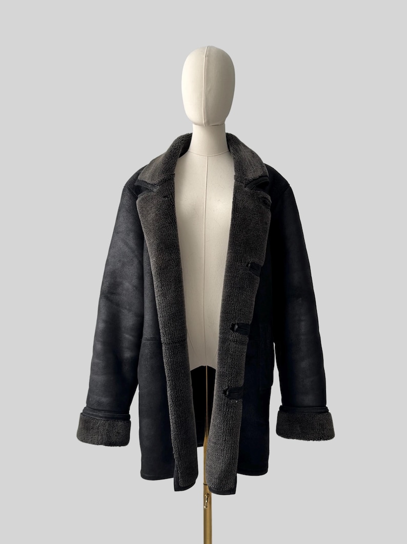 May include: A black shearling coat displayed on a mannequin. The coat features a dark gray, plush interior lining and collar, with matching cuffs. The exterior is a smooth, matte black. The coat has three dark buttons down the front.