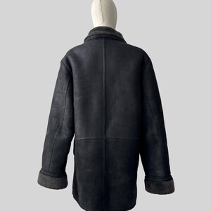 May include: Black shearling coat with a gray collar and cuffs. The coat is displayed on a mannequin. The coat has a classic design with a button closure and a tailored fit. The coat is made of a soft, supple material.