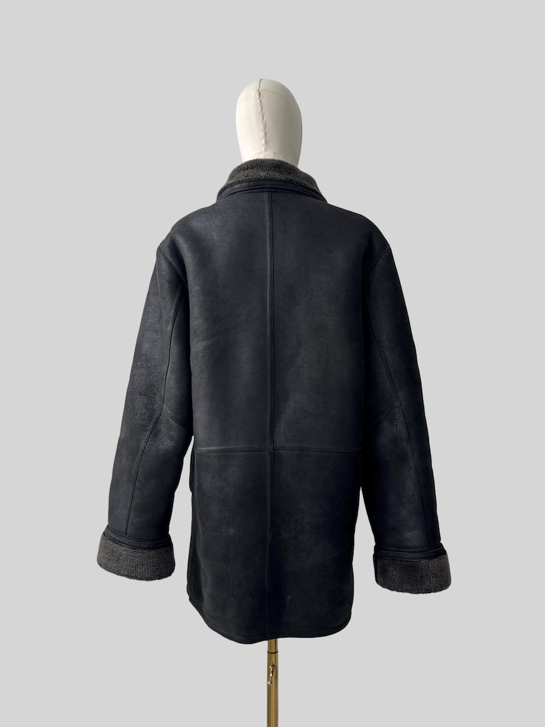 May include: Black shearling coat with a gray collar and cuffs. The coat is displayed on a mannequin. The coat has a classic design with a button closure and a tailored fit. The coat is made of a soft, supple material.