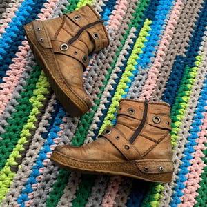 May include: A pair of brown leather ankle boots with buckles and metal eyelets. The boots have a zipper on the side and a thick platform sole. The boots are on a colorful crocheted rug.