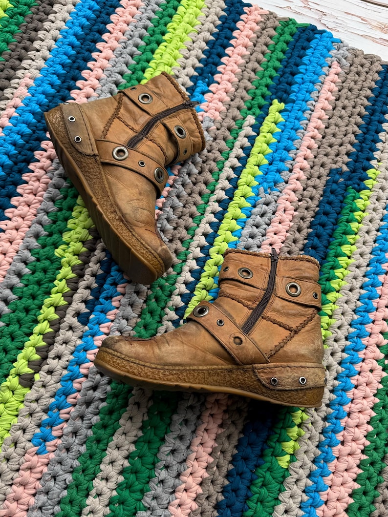 May include: A pair of brown leather ankle boots with buckles and metal eyelets. The boots have a zipper on the side and a thick platform sole. The boots are on a colorful crocheted rug.