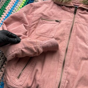May include: A pink corduroy jacket with a faux fur hood and a zipper closure. The jacket has a pocket on the left side and a ribbed hem.