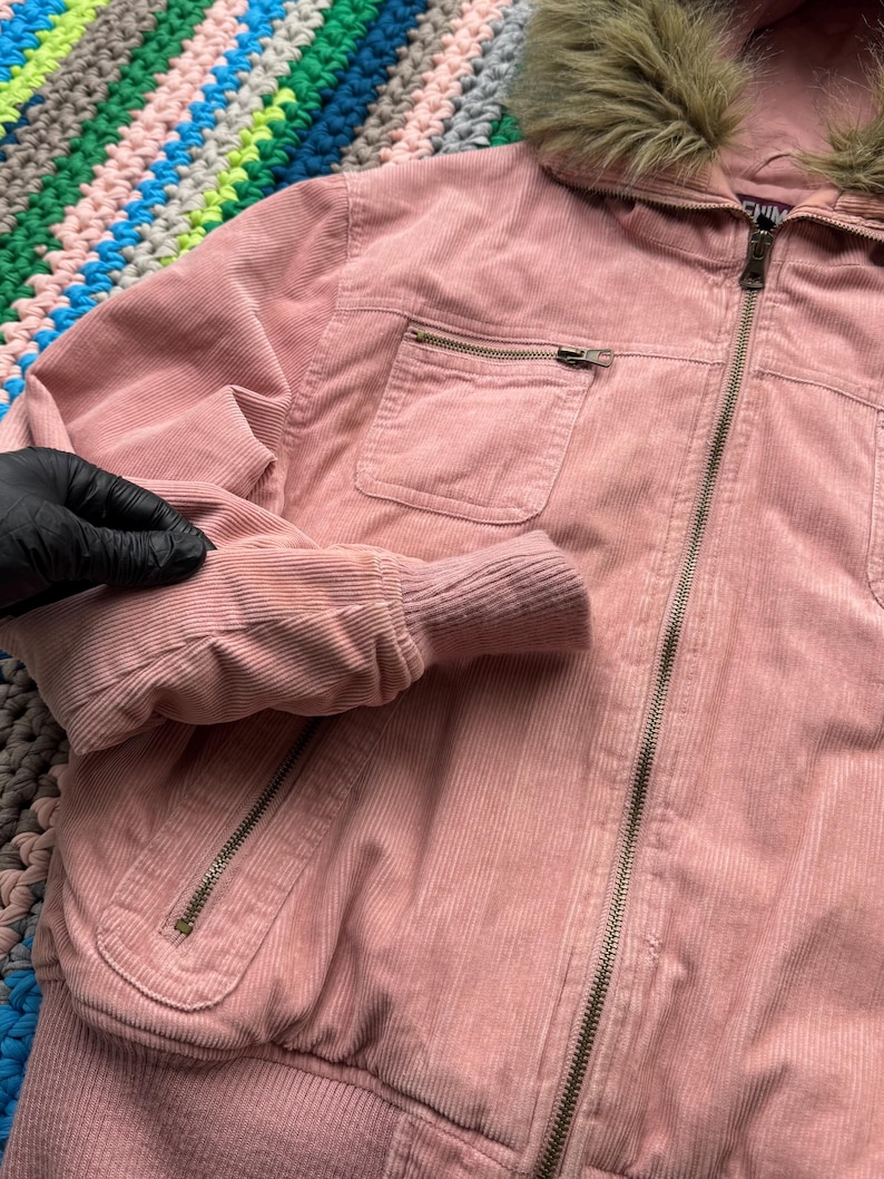 May include: A pink corduroy jacket with a faux fur hood and a zipper closure. The jacket has a pocket on the left side and a ribbed hem.