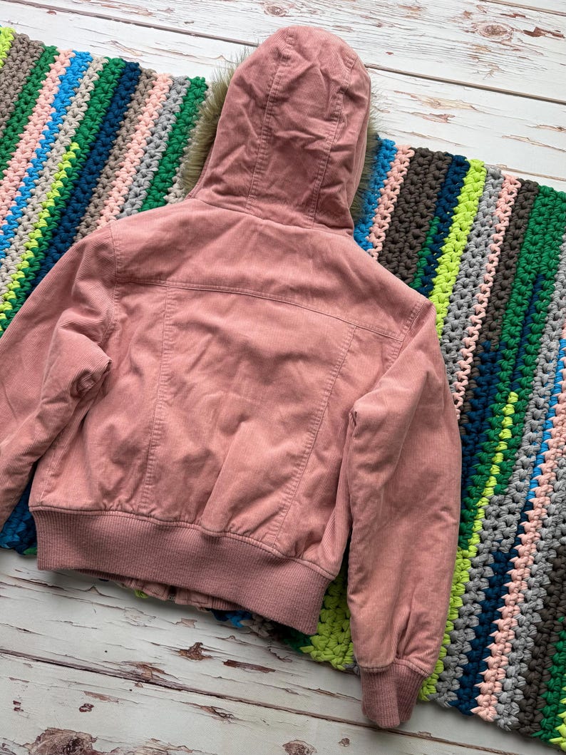 May include: A pink corduroy jacket with a faux fur lined hood. The jacket has a zipper closure and a ribbed waistband.