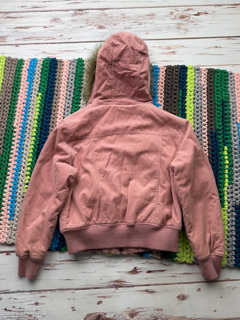 May include: A pink corduroy bomber jacket with a faux fur lined hood. The jacket has a ribbed hem and cuffs.
