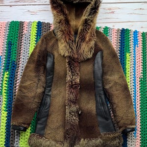 May include: A brown sheepskin coat with a fur-lined hood and black leather accents. The coat has a button closure and two pockets.