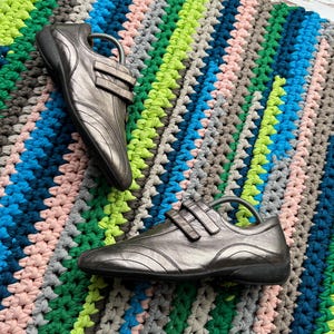 May include: A pair of silver leather shoes with velcro straps. The shoes are on a colorful crocheted rug.