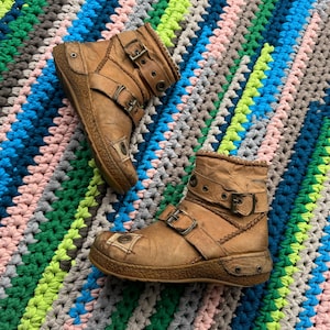 May include: A pair of brown leather ankle boots with multiple buckles and a distressed look. The boots are on a colorful crocheted rug.
