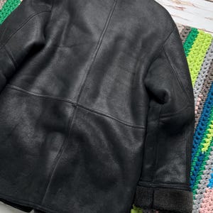 May include: A black leather jacket with a shearling collar and cuffs. The jacket has a classic design with visible stitching and a textured surface. The back of the jacket is shown, with a focus on the details of the leather.