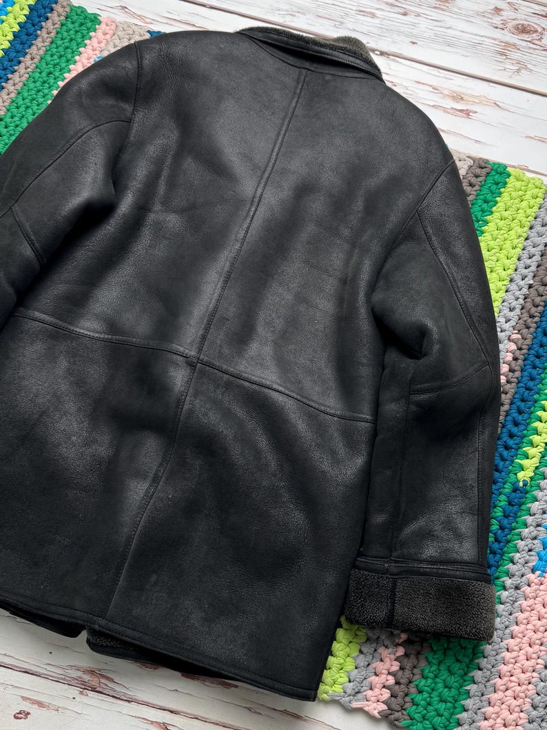 May include: A black leather jacket with a shearling collar and cuffs. The jacket has a classic design with visible stitching and a textured surface. The back of the jacket is shown, with a focus on the details of the leather.