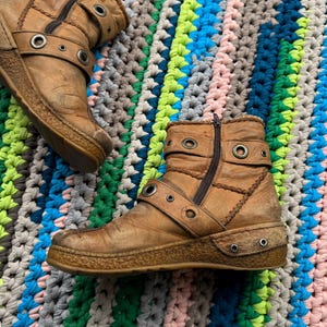 May include: A pair of brown leather ankle boots with buckles and metal eyelets. The boots have a zipper on the side and a worn look.