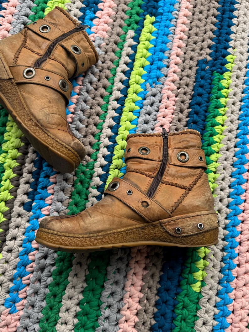 May include: A pair of brown leather ankle boots with buckles and metal eyelets. The boots have a zipper on the side and a worn look.