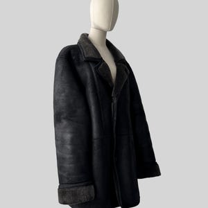 May include: A black shearling coat displayed on a mannequin. The coat features a classic lapel collar and turned-up cuffs with a contrasting dark gray shearling lining. The coat is buttoned and has a tailored fit, suggesting a vintage or classic style.