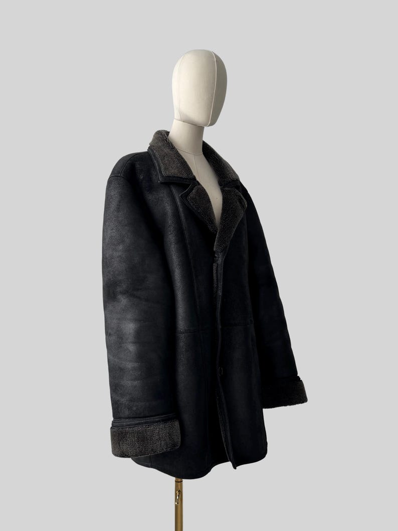 May include: A black shearling coat displayed on a mannequin. The coat features a classic lapel collar and turned-up cuffs with a contrasting dark gray shearling lining. The coat is buttoned and has a tailored fit, suggesting a vintage or classic style.