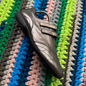 May include: A pair of silver leather shoes with a strap and buckle closure. The shoes are on a colorful crocheted surface.