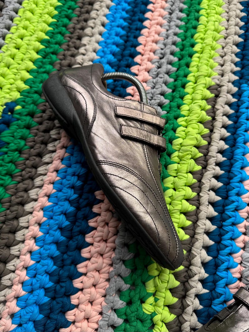 May include: A pair of silver leather shoes with a strap and buckle closure. The shoes are on a colorful crocheted surface.