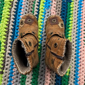 May include: A pair of brown leather boots with zipper closures and metal accents. The boots have a distressed look and are lined with fur. The boots are photographed on a colorful crocheted rug.