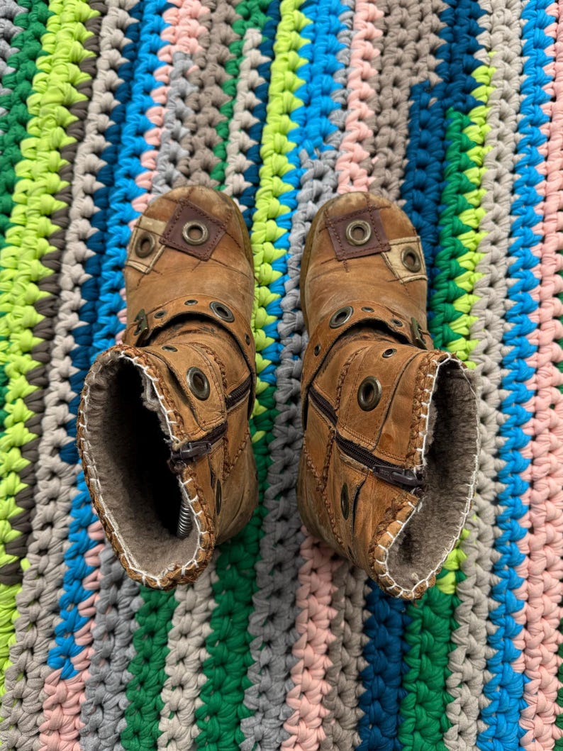 May include: A pair of brown leather boots with zipper closures and metal accents. The boots have a distressed look and are lined with fur. The boots are photographed on a colorful crocheted rug.