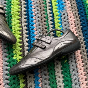 May include: A pair of silver leather shoes with two straps across the top. The shoes are on a colorful crocheted rug.