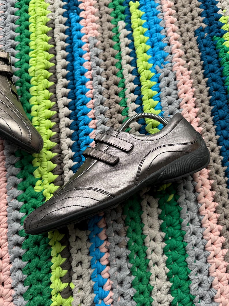 May include: A pair of silver leather shoes with two straps across the top. The shoes are on a colorful crocheted rug.
