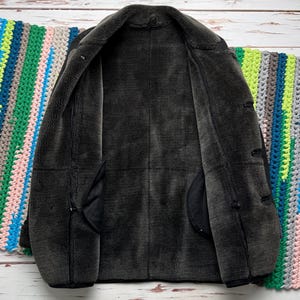 May include: A dark charcoal gray jacket with a thick, plush, dark gray faux fur lining. The jacket features a wide collar, long sleeves, and a front opening with visible toggle-style closures along the side, showcasing a soft, textured interior.