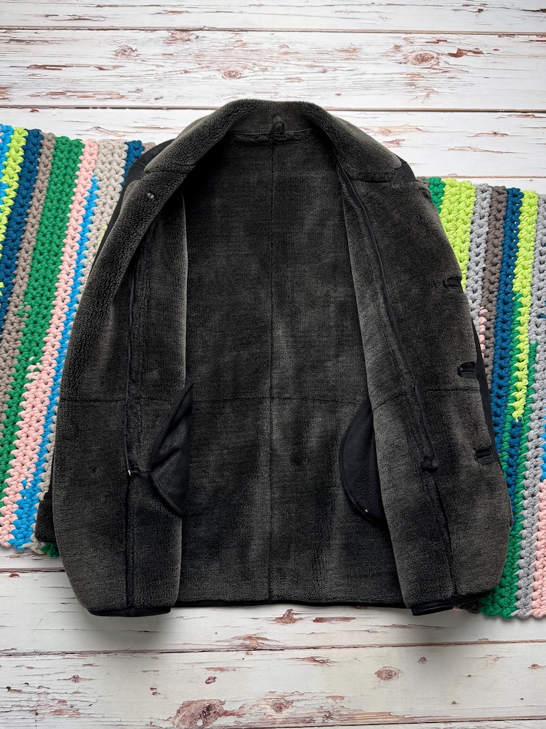 May include: A dark charcoal gray jacket with a thick, plush, dark gray faux fur lining. The jacket features a wide collar, long sleeves, and a front opening with visible toggle-style closures along the side, showcasing a soft, textured interior.
