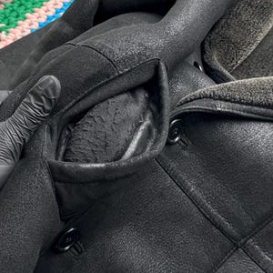 May include: Close-up of a black leather coat with a shearling collar and lining. The coat features a double-breasted design with large black buttons and a textured finish. The pocket lining is visible, and the coat appears to be well-worn.