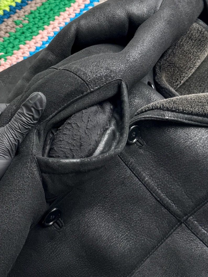 May include: Close-up of a black leather coat with a shearling collar and lining. The coat features a double-breasted design with large black buttons and a textured finish. The pocket lining is visible, and the coat appears to be well-worn.