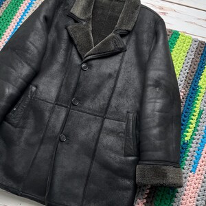 May include: A black leather coat with a shearling collar and lining. The coat has a button-up front, two front pockets, and shearling trim on the cuffs. The coat is displayed on a colorful crocheted blanket.