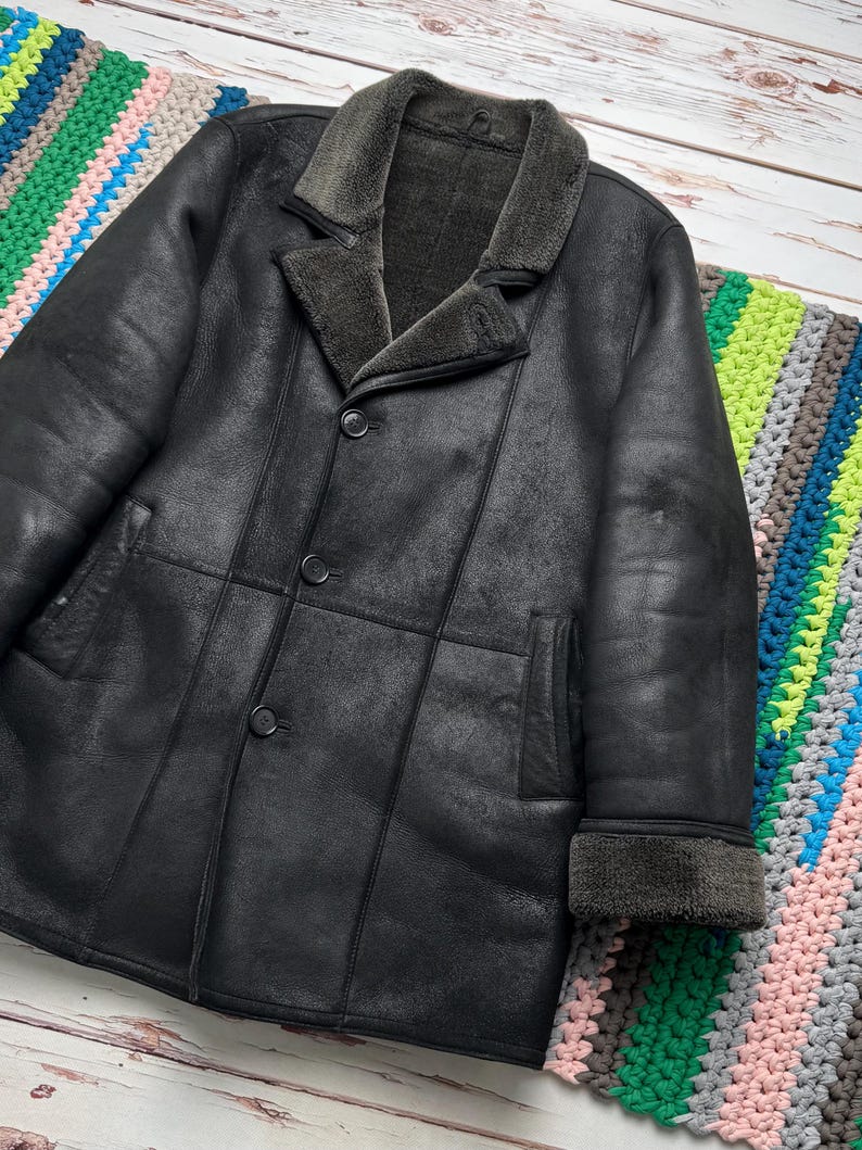May include: A black leather coat with a shearling collar and lining. The coat has a button-up front, two front pockets, and shearling trim on the cuffs. The coat is displayed on a colorful crocheted blanket.