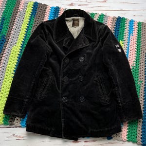 May include: A black corduroy pea coat featuring a double-breasted front with six buttons, wide lapels, and two side pockets. The jacket includes a light-colored interior lining and a small circular patch on the left sleeve with the letters GS.