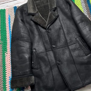 May include: A black leather coat with a shearling collar and lining. The coat has a button-up front, two front pockets, and a classic tailored design. The coat is displayed on a colorful, striped textile.