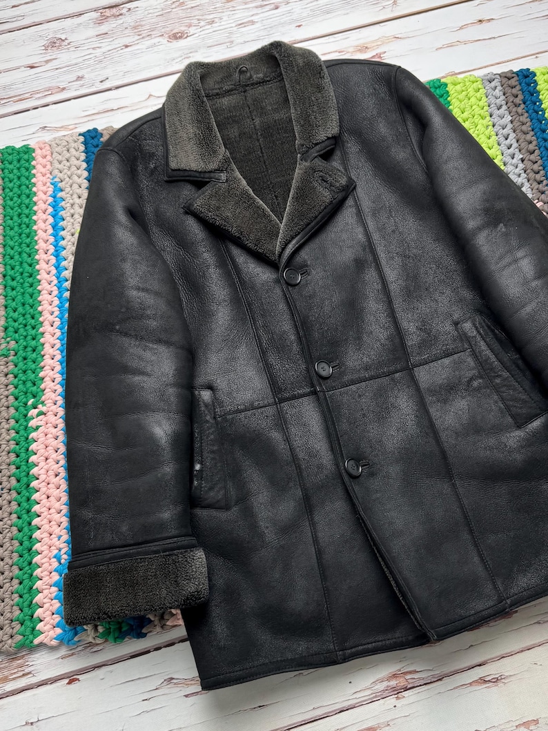 May include: A black leather coat with a shearling collar and lining. The coat has a button-up front, two front pockets, and a classic tailored design. The coat is displayed on a colorful, striped textile.