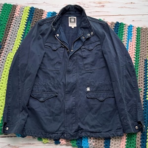May include: A dark navy blue G-Star RAW field jacket featuring a high collar with snap buttons, a front zipper closure, and four front pockets with flaps. The jacket includes a small white brand logo on the lower left pocket and long sleeves.