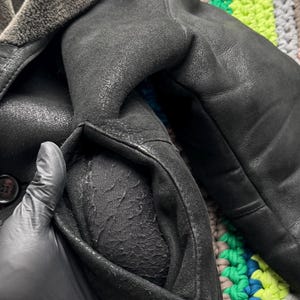 May include: Close-up of a black leather coat with a shearling collar. The coat has a classic design with a button closure and a pocket. The leather appears worn, giving it a vintage look. The coat is likely a winter garment.