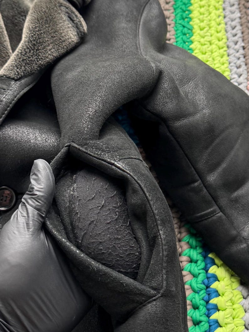 May include: Close-up of a black leather coat with a shearling collar. The coat has a classic design with a button closure and a pocket. The leather appears worn, giving it a vintage look. The coat is likely a winter garment.