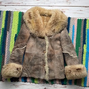May include: A brown suede coat with a large, fluffy, light brown fur collar and cuffs. The coat has a button-up front and decorative stitching along the seams. The coat is displayed on a colorful, striped crocheted blanket.