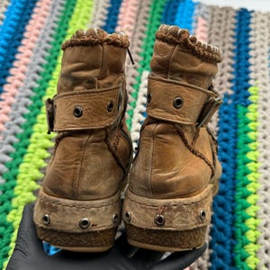 May include: A pair of brown leather ankle boots with a decorative stitching detail around the top of the boot. The boots have a buckle closure and a thick, textured sole.