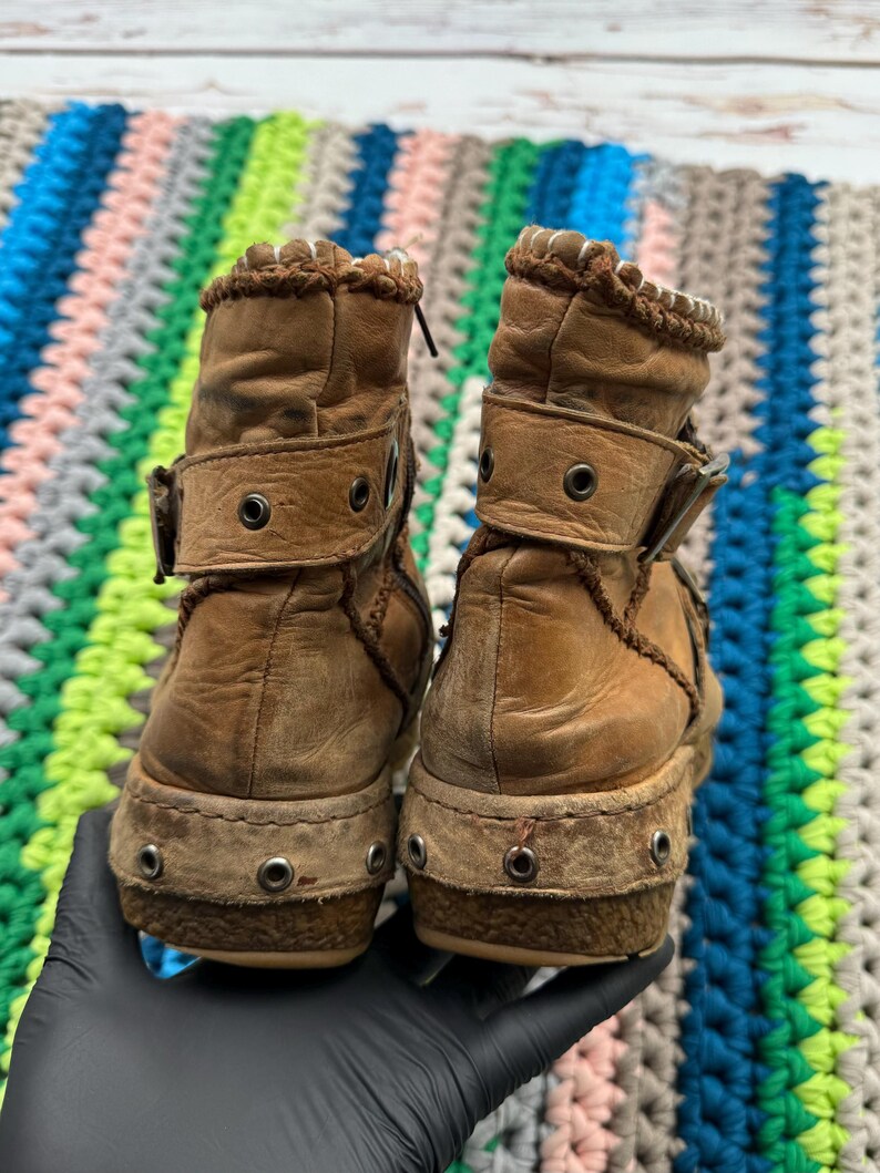 May include: A pair of brown leather ankle boots with a decorative stitching detail around the top of the boot. The boots have a buckle closure and a thick, textured sole.