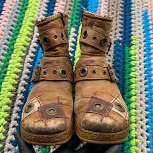 May include: A pair of brown leather ankle boots with multiple buckles and decorative stitching. The boots have a distressed look and are made of a patchwork of different materials.