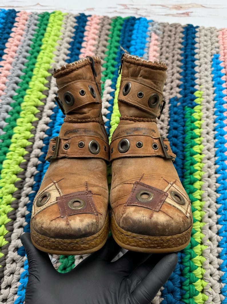 May include: A pair of brown leather ankle boots with multiple buckles and decorative stitching. The boots have a distressed look and are made of a patchwork of different materials.