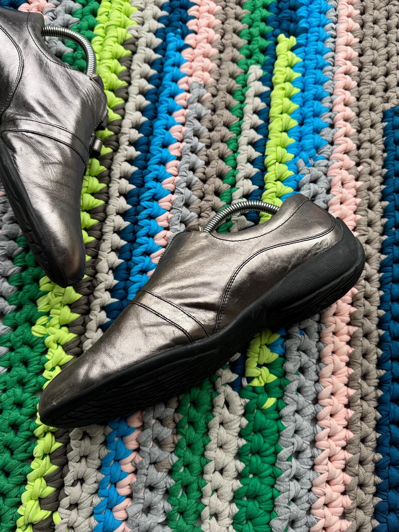 May include: A pair of silver leather shoes with black soles. The shoes are on a colorful crocheted rug.