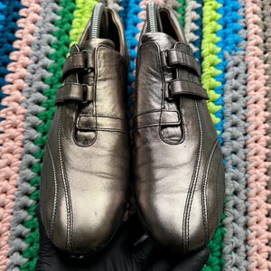 May include: A pair of silver leather shoes with a buckle closure. The shoes have a sporty design with a stitched detail on the sides.