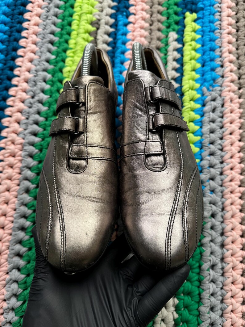May include: A pair of silver leather shoes with a buckle closure. The shoes have a sporty design with a stitched detail on the sides.
