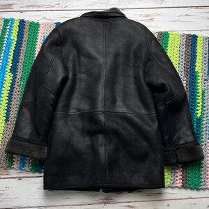 May include: A black, faux-leather coat with a shearling-style collar and cuffs. The coat has a classic design with a button closure and a tailored fit. The coat is displayed on a wooden surface with a colorful blanket.