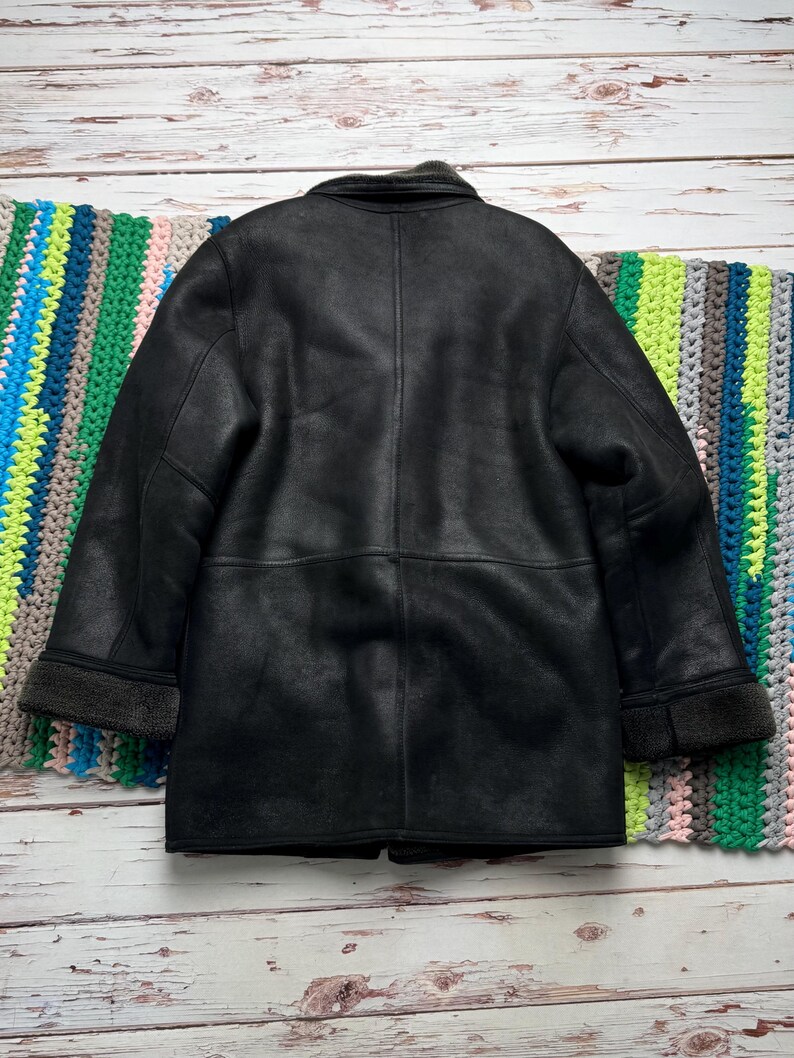 May include: A black, faux-leather coat with a shearling-style collar and cuffs. The coat has a classic design with a button closure and a tailored fit. The coat is displayed on a wooden surface with a colorful blanket.